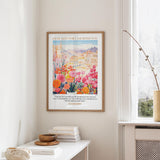 a framed poster of a colorful floral scene hanging on a wall, with a stack of books and a vase on a table below it.
