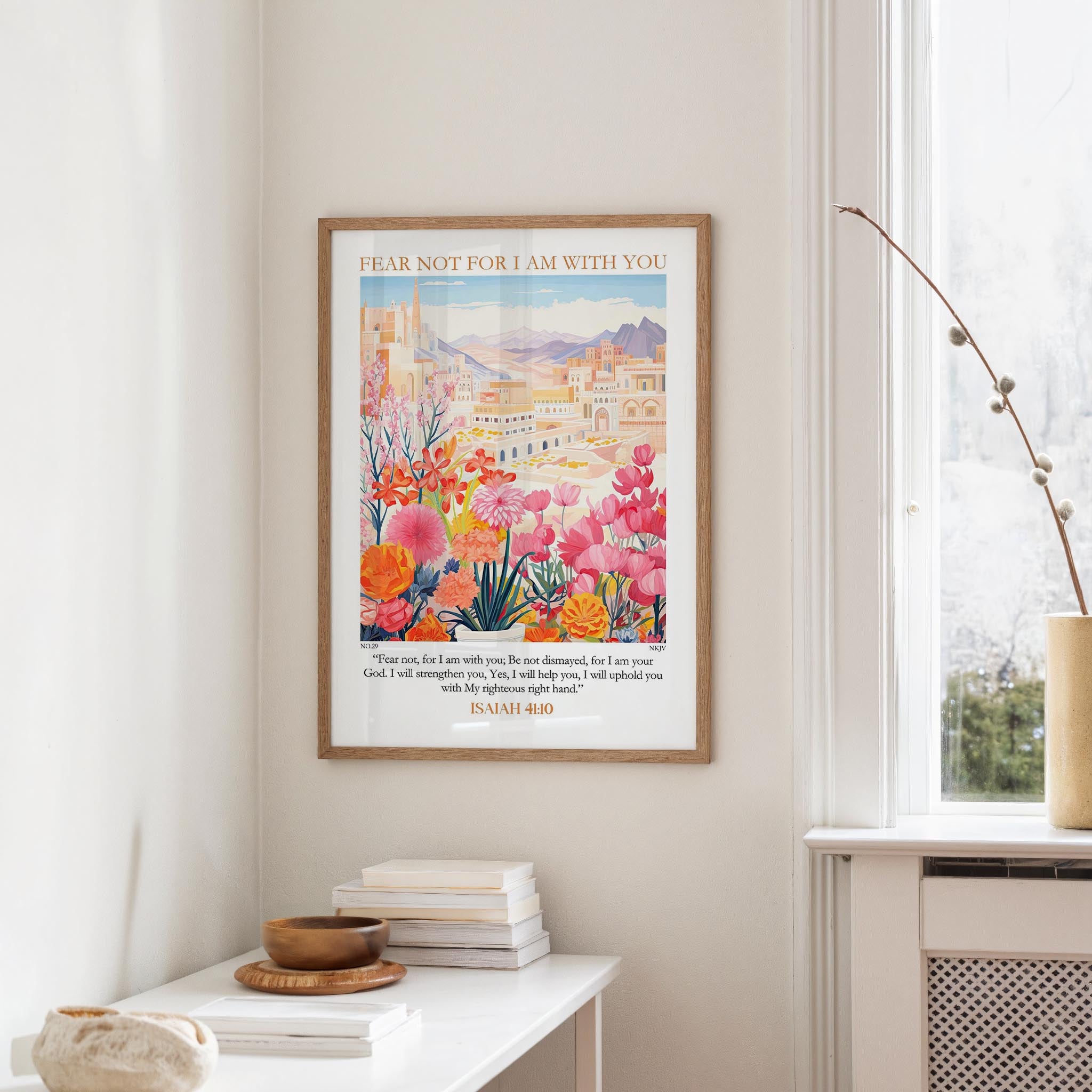 a framed poster of a colorful floral scene hanging on a wall, with a stack of books and a vase on a table below it.