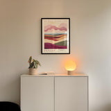 a minimalist interior design with a framed poster of a landscape, a potted plant, and a small lamp on a white cabinet.