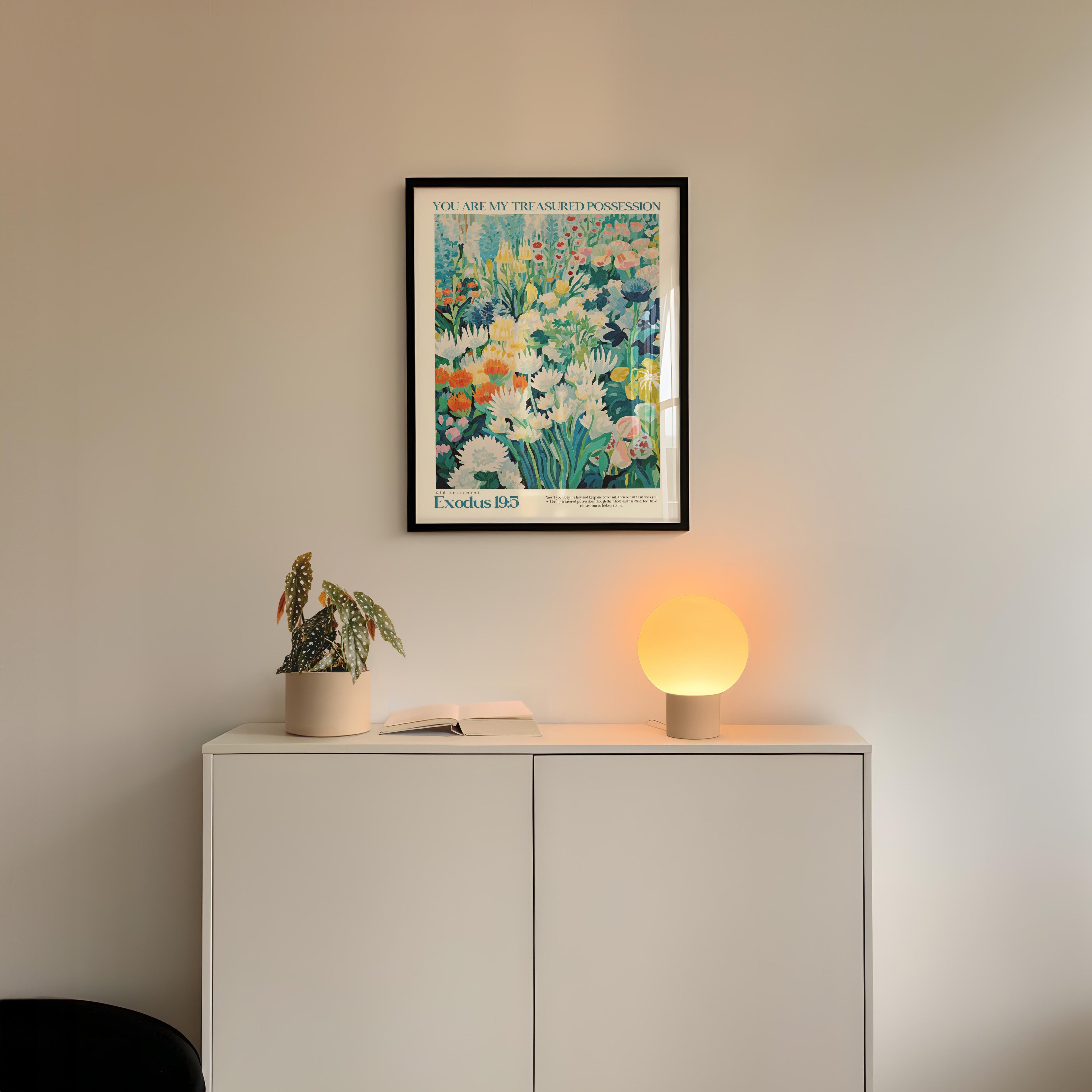 a white cabinet with a potted plant and a lamp on top, set against a white wall. Above the cabinet, there is a framed poster with a colorful floral design.
