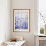 a framed poster of a serene landscape with a person standing in the foreground, surrounded by a field of purple flowers. The poster is hung on a wall above a white table, which has a stack of books and a small bowl on it.