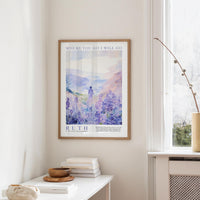 a framed poster of a serene landscape with a person standing in the foreground, surrounded by a field of purple flowers. The poster is hung on a wall above a white table, which has a stack of books and a small bowl on it.