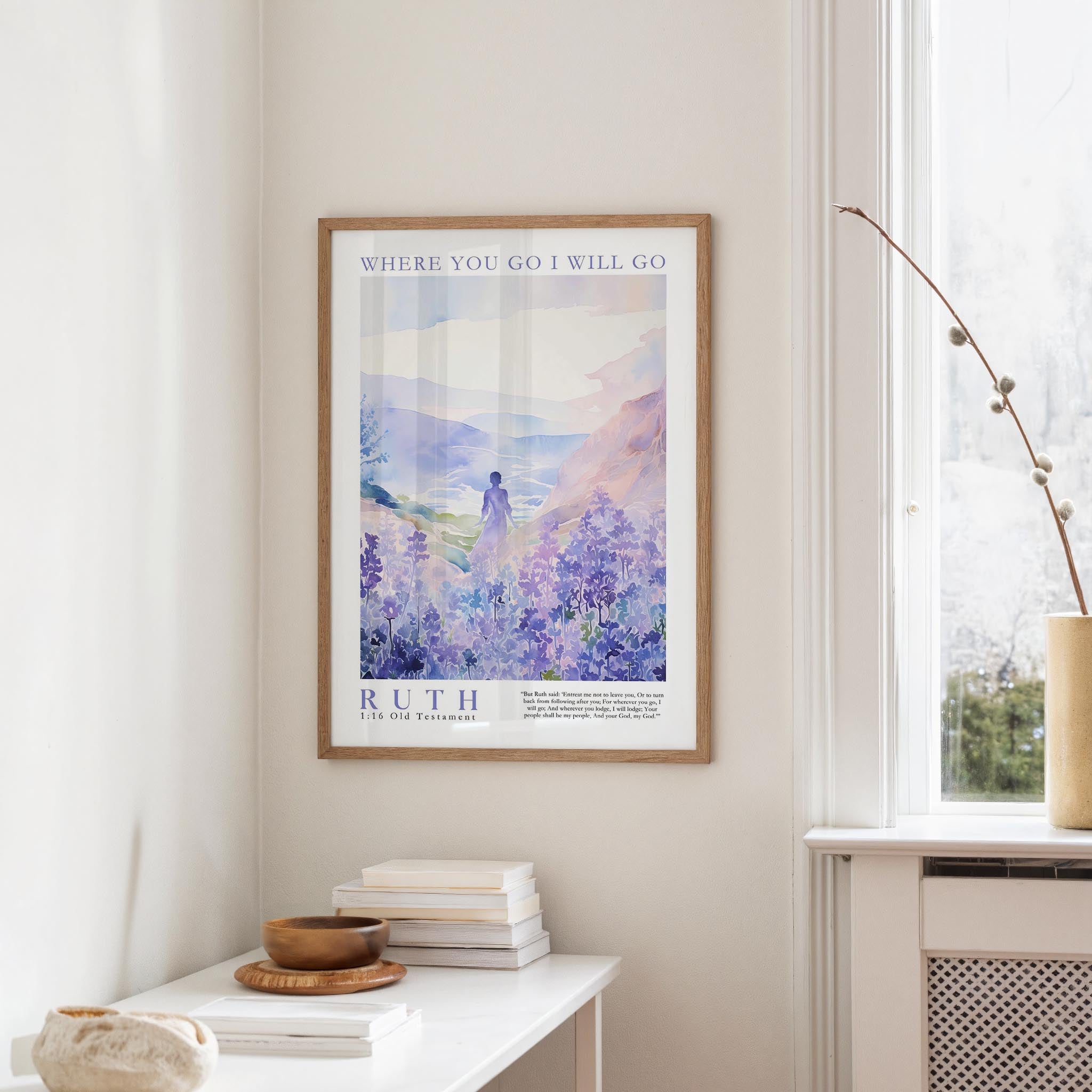 a framed poster of a serene landscape with a person standing in the foreground, surrounded by a field of purple flowers. The poster is hung on a wall above a white table, which has a stack of books and a small bowl on it.
