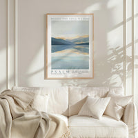 a framed poster of a serene landscape with mountains and a lake, hanging on a white wall above a white couch with a blanket draped over it.