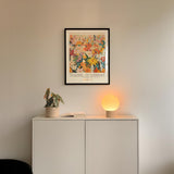 a white cabinet with a potted plant and a lamp on top, set against a white wall. Above the cabinet, there is a framed poster with a colorful floral design.