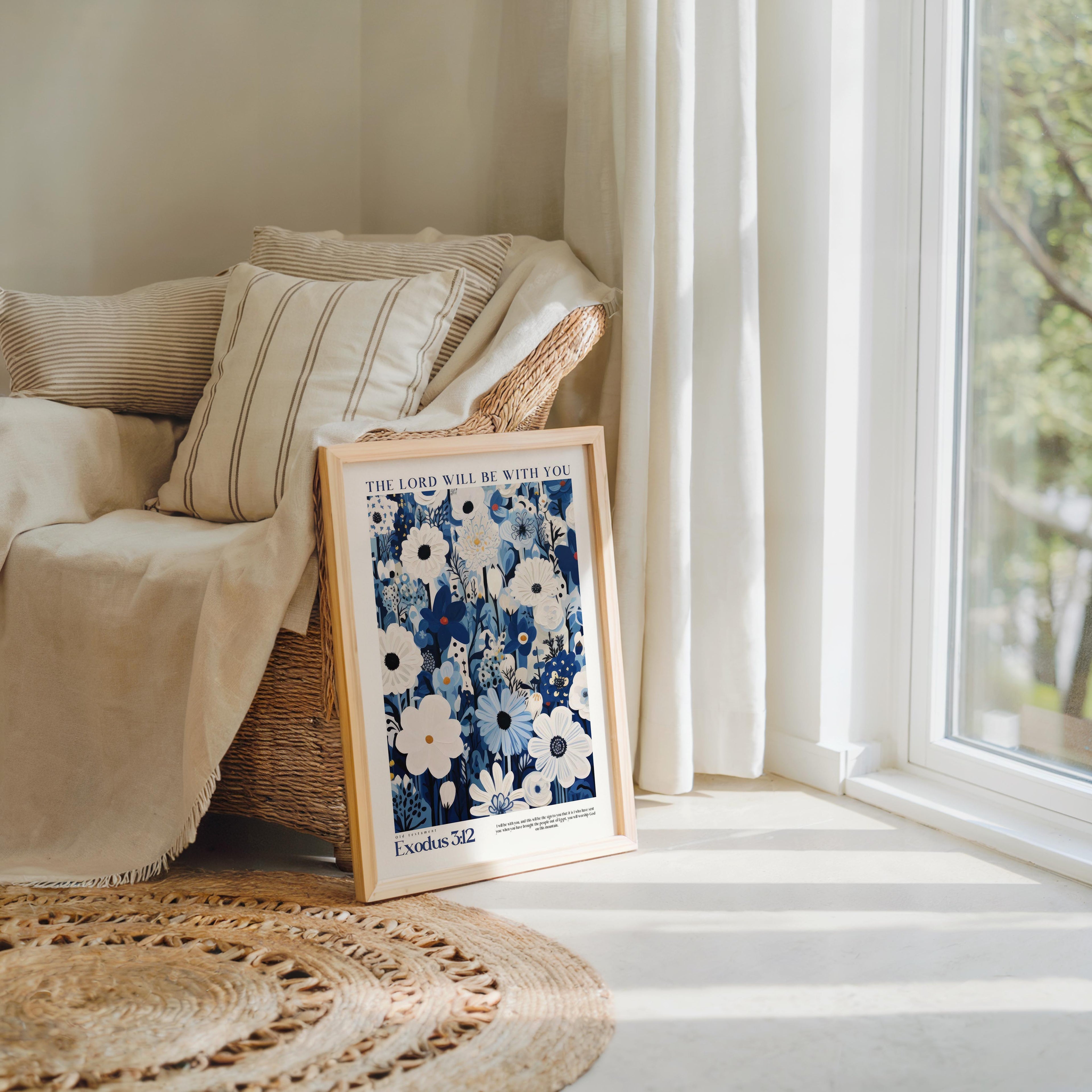 A framed poster with a floral design is propped up against a beige armchair in a cozy living room, with a large window and a woven rug on the floor.