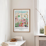 a framed poster of a colorful garden scene hanging on a wall, with a stack of books and a vase on a table below it.