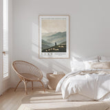 A bedroom with a large bed, a woven chair, and a framed poster of a pastoral scene hanging on the wall.