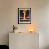 a white cabinet with a plant and a lamp on top, and a framed poster of a sunset above it.