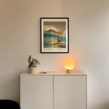 a minimalist interior design with a framed poster of a landscape, a potted plant, and a small lamp on a white cabinet.