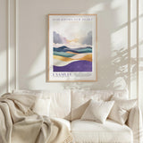a framed poster of a landscape with mountains and a quote from the Bible, "I Samuel", hanging on a white wall above a white couch with a blanket draped over it.