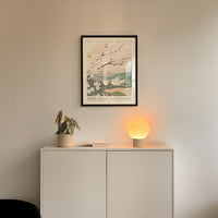 a minimalist interior design with a framed poster of a floral scene hanging on a white wall above a white cabinet with a plant and a lamp on top.