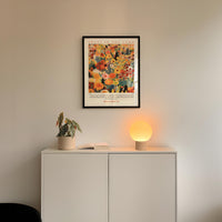 a white cabinet with a potted plant and a lamp on top, set against a white wall. Above the cabinet, there is a framed poster with a colorful floral design.