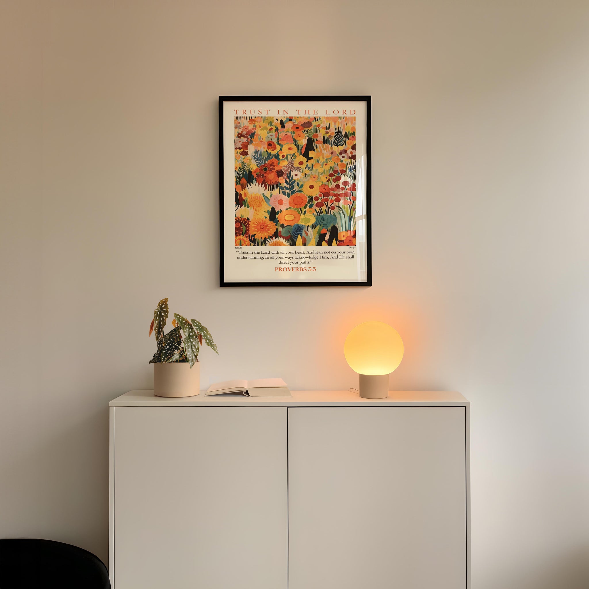 a white cabinet with a potted plant and a lamp on top, set against a white wall. Above the cabinet, there is a framed poster with a colorful floral design.