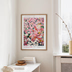 a framed poster of a colorful floral arrangement hanging on a wall, with a white table and a vase of flowers nearby.