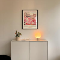a white cabinet with a potted plant and a lamp on top, set against a white wall. Above the cabinet, there is a framed poster with a floral design.