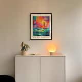 a white cabinet with a potted plant and a lamp on top, and a framed poster of a colorful abstract design hanging on the wall above the cabinet.