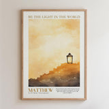 The image is a framed poster with a watercolor painting of a lighthouse on a hill, set against a cloudy sky. The text on the poster reads "BE THE LIGHT IN THE WORLD" and "MATTHEW 3:13 New Testament".