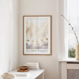 a framed poster of a serene landscape with clouds and crowns, hanging on a wall in a room with a white table and a window.