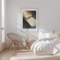 a serene bedroom scene with a bed, a woven chair, and a framed poster of a roll of paper on the wall.