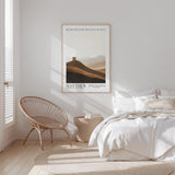 A minimalist bedroom with a bed, a woven chair, and a framed poster of a mountain landscape.