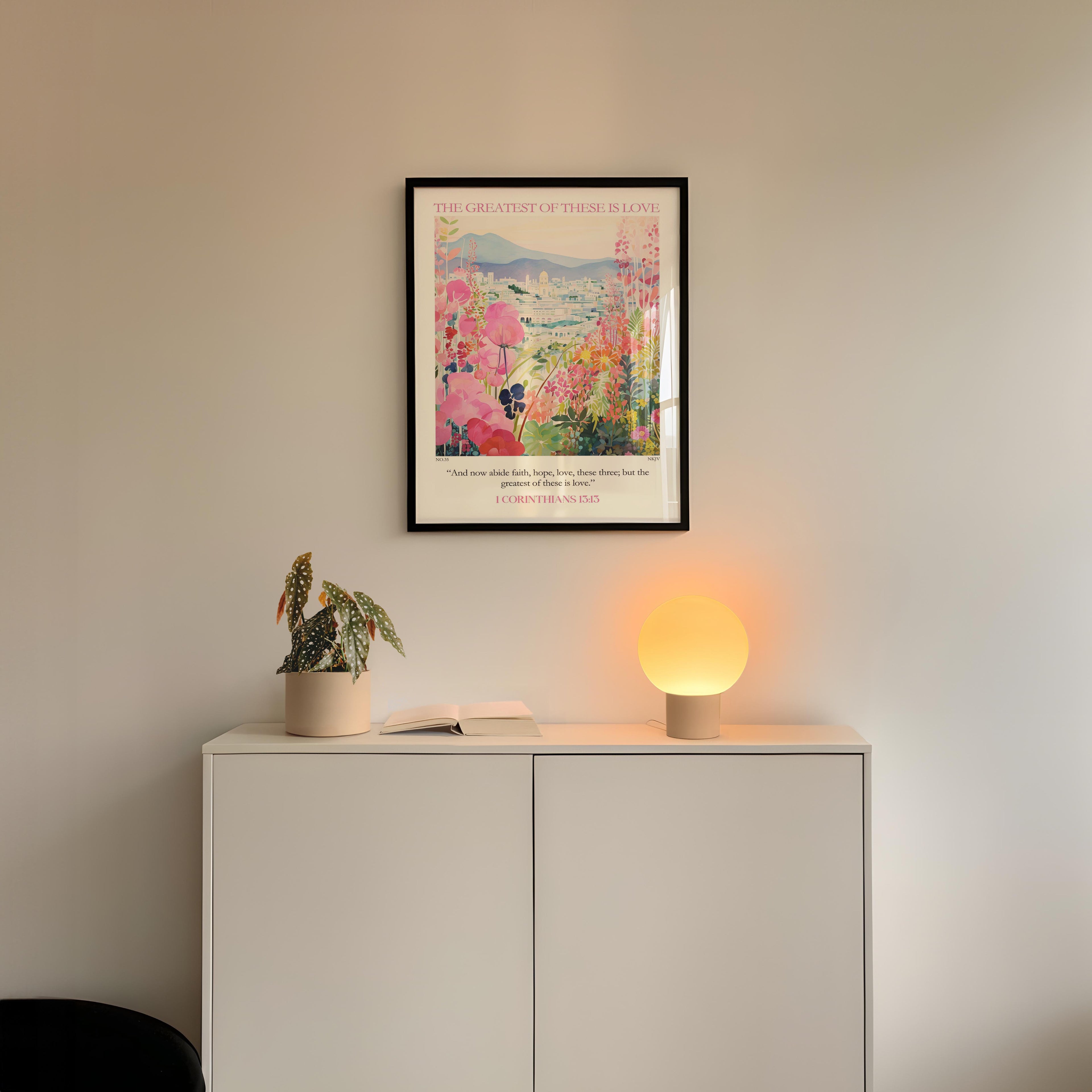 a white cabinet with a potted plant and a lamp on top, set against a white wall. Above the cabinet, there is a framed poster with a floral design.