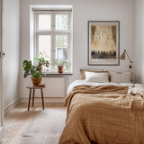 a cozy bedroom with a large bed, a nightstand, and a potted plant. The room has a neutral color scheme with white walls and a wooden floor. A framed poster is hanging on the wall above the bed, and a window with a plant on the sill is visible.