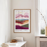 a framed poster of a landscape with mountains and hills, hanging on a wall in a room with a window and a small table with books and a bowl on it.