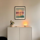a minimalist interior design with a framed poster of a sunset on the wall above a white cabinet with a plant and a lamp on top.