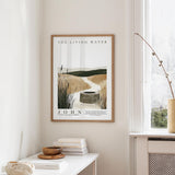 a framed poster of a serene landscape with a well in the center, hanging on a wall above a white table with a stack of books and a small vase.