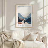 a framed poster of a mountain landscape with a person walking through it, hanging on a wall above a white couch with a blanket draped over it.
