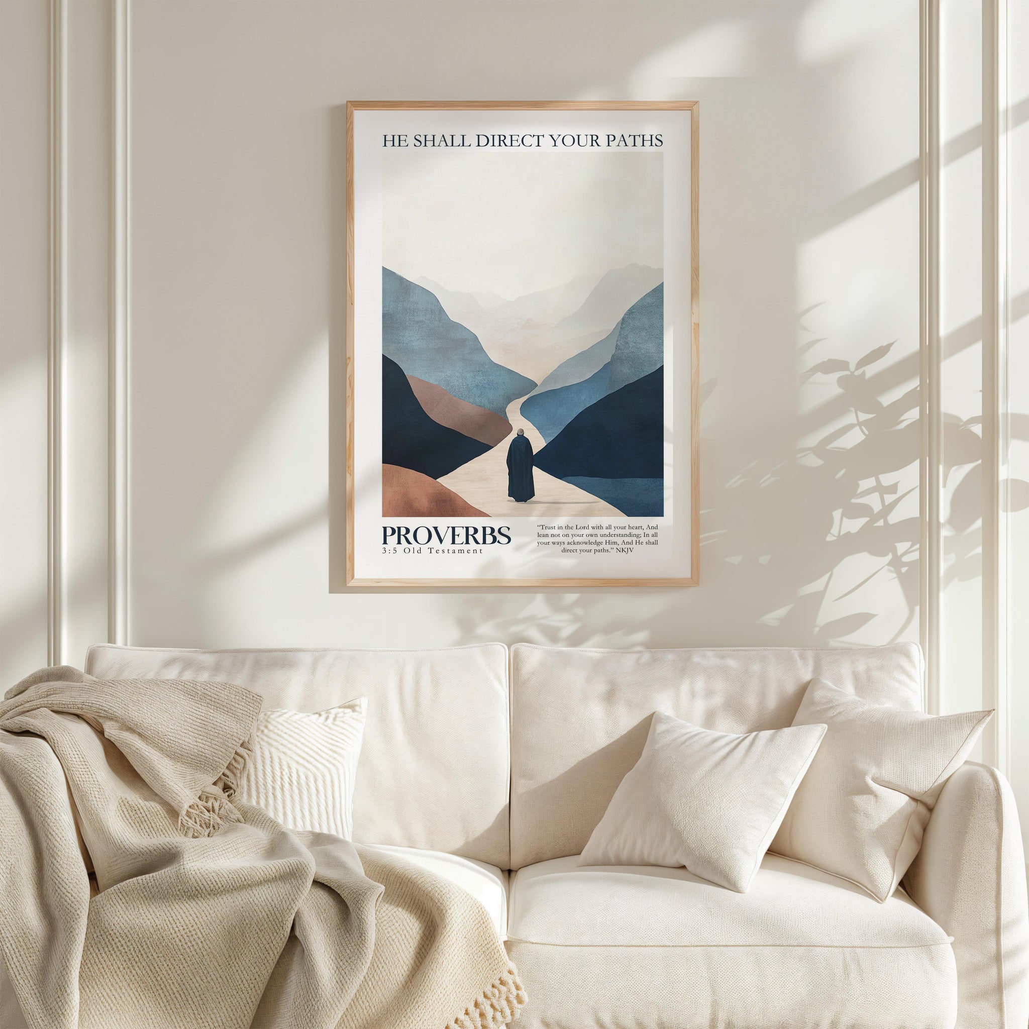 a framed poster of a mountain landscape with a person walking through it, hanging on a wall above a white couch with a blanket draped over it.