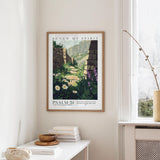 a framed poster of a scenic landscape with a stone wall and flowers hanging on a wall, along with a window and a small table with books and a bowl on it.