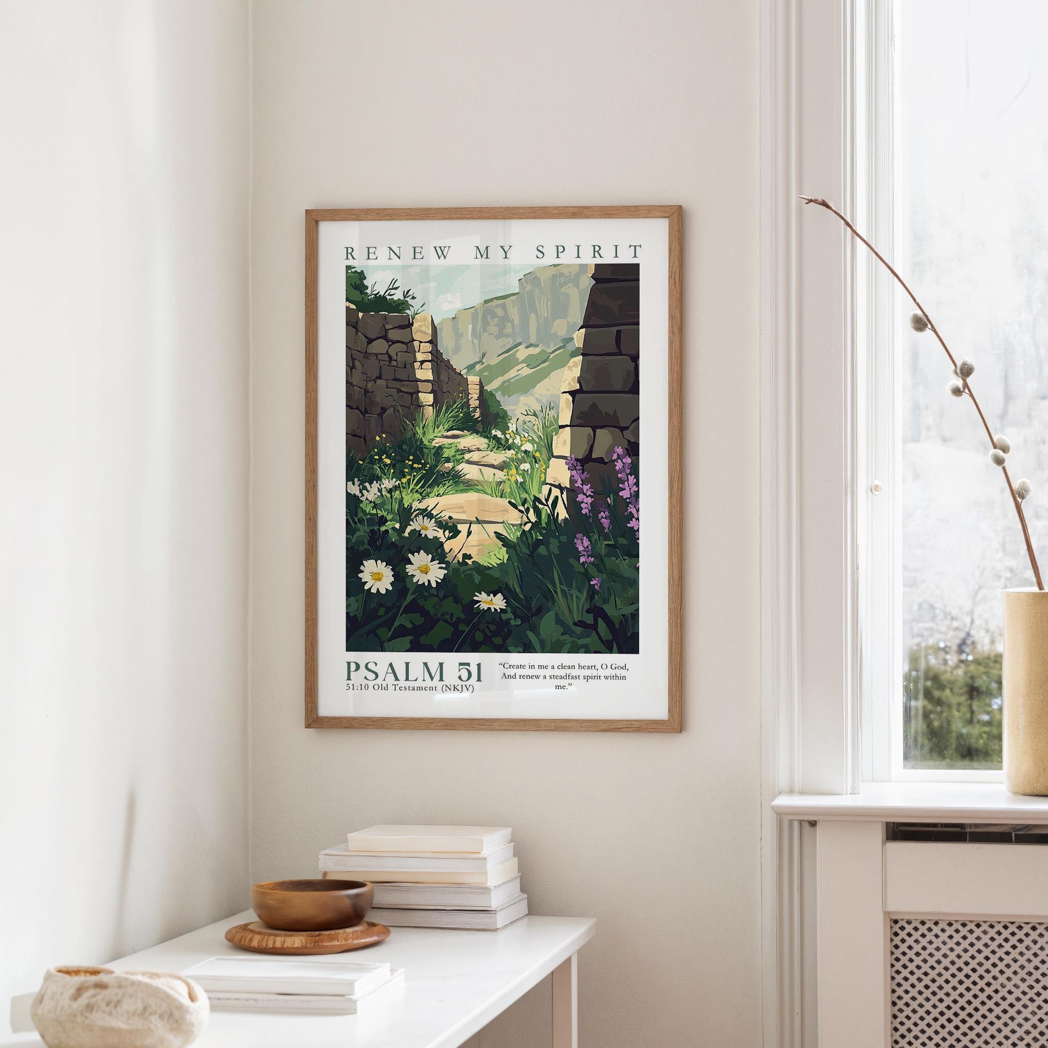 a framed poster of a scenic landscape with a stone wall and flowers hanging on a wall, along with a window and a small table with books and a bowl on it.