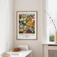 a framed poster of a colorful floral arrangement hanging on a wall, with a window and a small table with books and a bowl in the foreground.