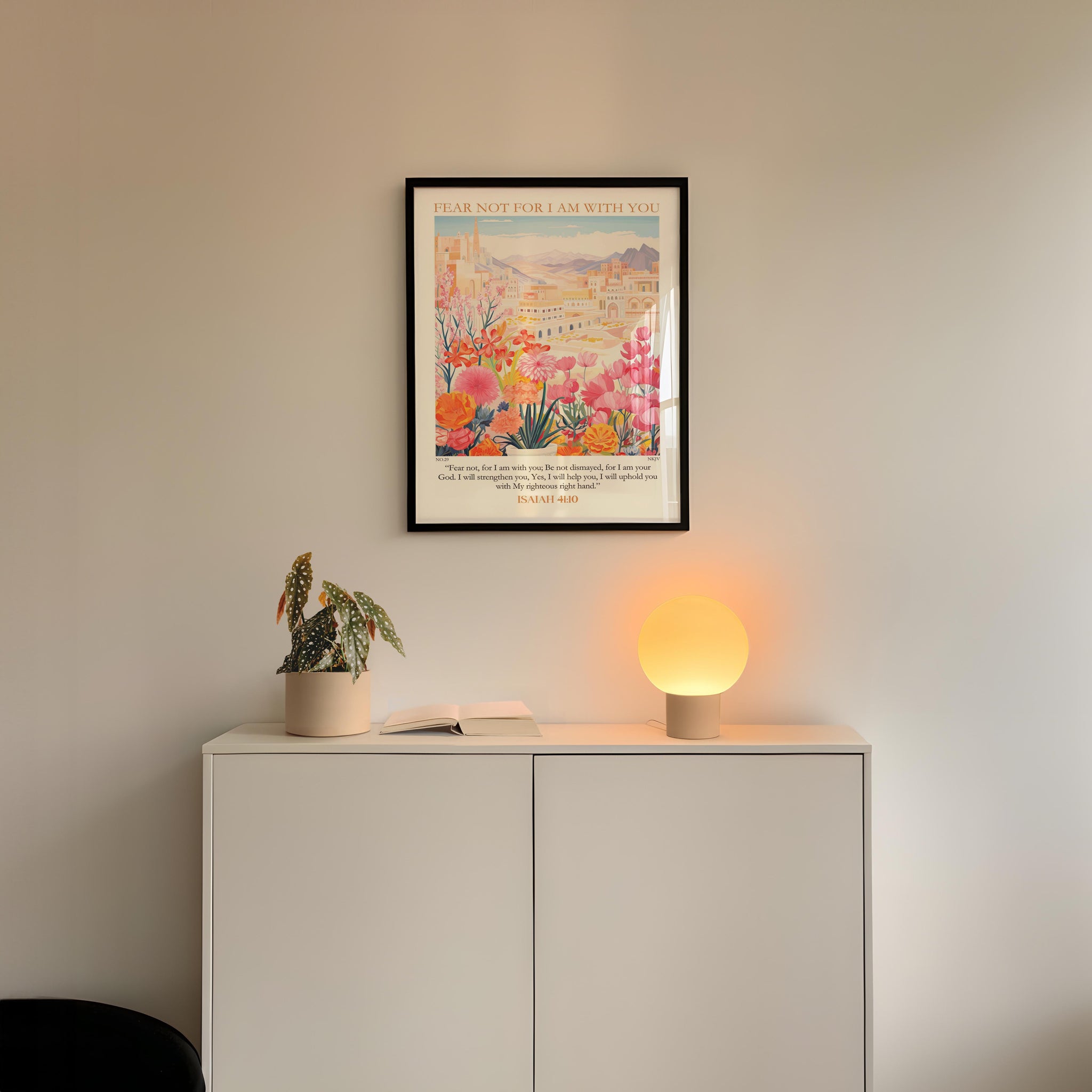 a white cabinet with a potted plant and a lamp on top, set against a white wall. Above the cabinet, there is a framed poster with a colorful illustration of a cityscape.