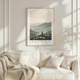 a framed poster of a pastoral scene with a shepherd and his flock of sheep, hanging on a wall above a white couch with a blanket draped over it.
