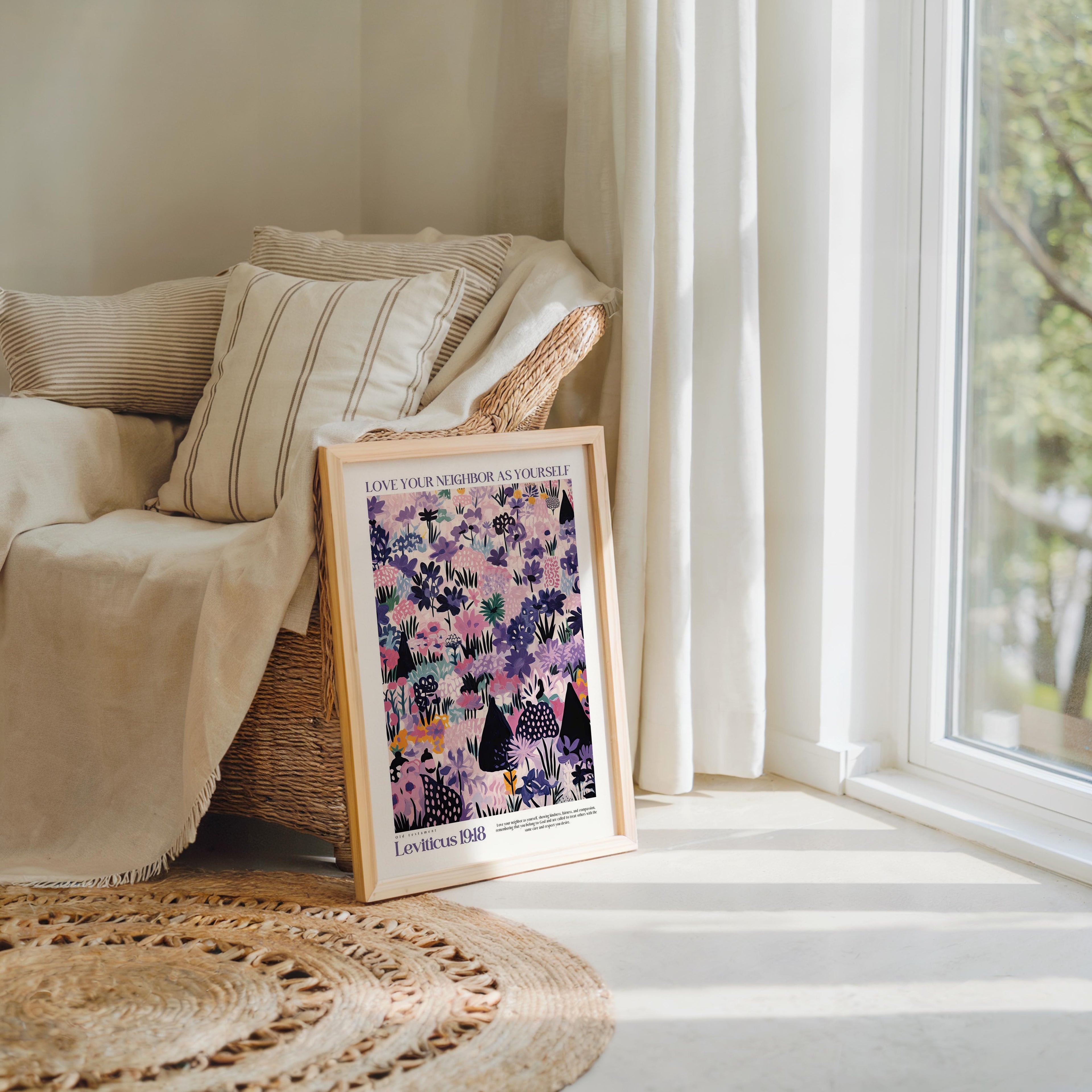 A framed poster with a colorful floral design is propped up against a beige sofa in a cozy living room, with a large window and a woven rug visible in the background.