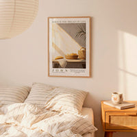 a bedroom scene with a framed poster of a loaf of bread and a basket of fruit hanging on the wall above a bed with striped bedding.