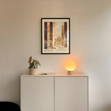 a minimalist interior design with a framed poster of a building and a small potted plant on a white cabinet. The poster has the text "GENESIS" and "God can turn evil into good". The cabinet has a small lamp on top, and there is a book on the shelf below the lamp.