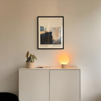 a minimalist interior design with a framed poster of a mountain landscape above a white cabinet with a plant and a lamp on top.
