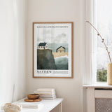 a framed poster of a coastal scene with houses and cliffs, hanging on a wall in a room with a white table and a window.