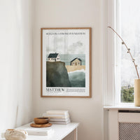a framed poster of a coastal scene with houses and cliffs, hanging on a wall in a room with a white table and a window.