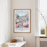 a framed poster of a colorful floral landscape hanging on a wall, with a window and a small table with books and a bowl in the foreground.