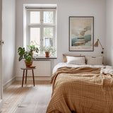 A cozy bedroom with a large bed, a nightstand, and a potted plant. The room has a neutral color scheme with white walls and a wooden floor. A framed poster of a staircase is displayed on the wall above the bed.