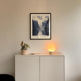 a minimalist interior design with a framed poster of a mountain landscape hanging on a white wall above a white cabinet. The cabinet has a plant and a lamp on it, creating a cozy and inviting atmosphere.