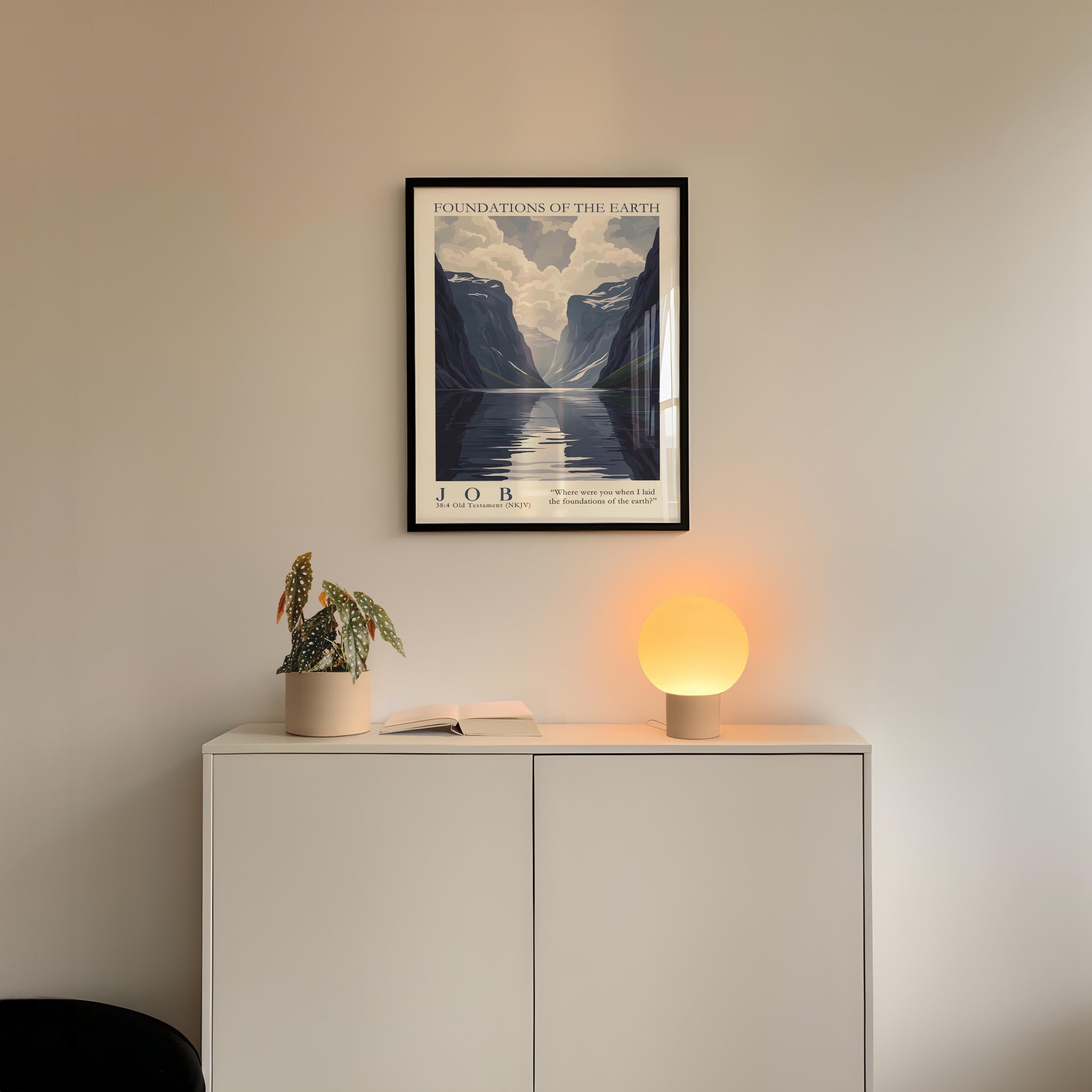 a minimalist interior design with a framed poster of a mountain landscape hanging on a white wall above a white cabinet. The cabinet has a plant and a lamp on it, creating a cozy and inviting atmosphere.