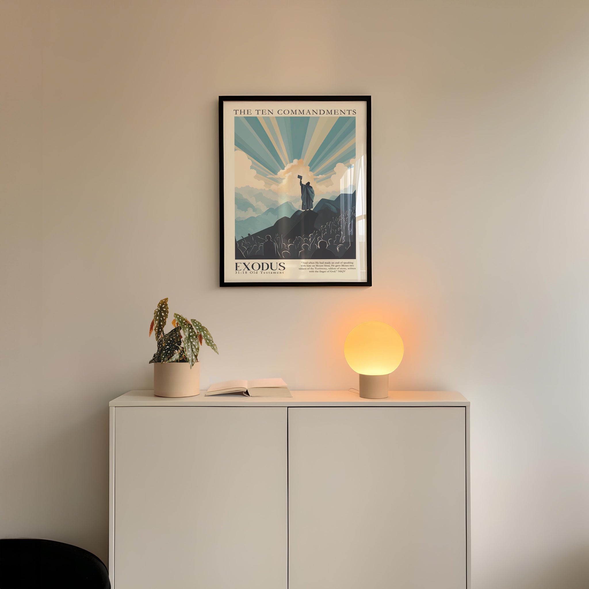 a framed poster of the Ten Commandments hanging on a wall above a white cabinet with a plant and a lamp on top.