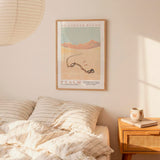 a bedroom scene with a bed, a nightstand, and a poster on the wall. The poster is a framed artwork depicting a desert landscape with mountains in the background.