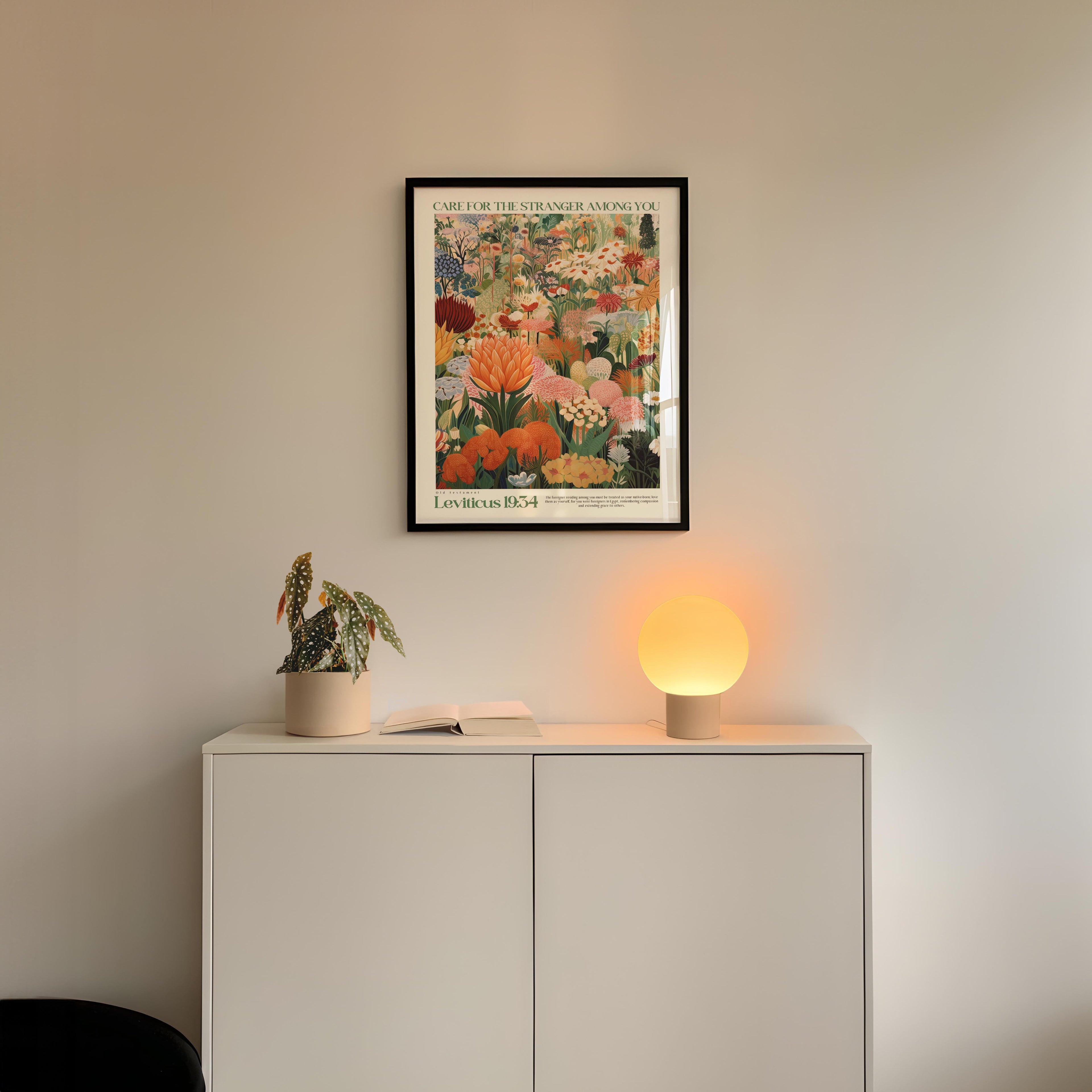 a white cabinet with a potted plant and a lamp on top, set against a white wall. Above the cabinet, there is a framed poster with a colorful floral design.